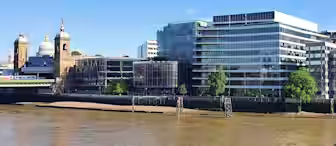 A view of modern and historical buildings lining the River Thames in London, featuring contemporary glass structures alongside traditional architecture with domes. The sky is clear and blue, reflecting on the water below.