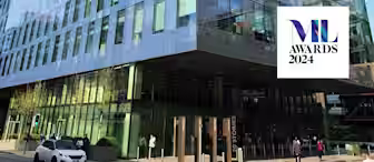 A modern glass building with multiple reflections and light coming in from different angles. A sign to the right reads "ML Awards 2024". The scene is lively, with several pedestrians walking and a white car parked in front.
