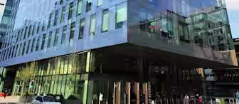 A tall, modern glass office building with a reflective facade stands in an urban area under a blue sky with scattered clouds. A few pedestrians can be seen near the entrance, and a white car is parked nearby.
