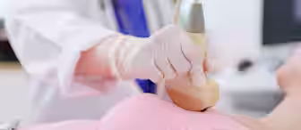 A healthcare professional wearing gloves performs an ultrasound scan on a patient’s breast using a handheld device. The patient is lying down wearing a pink top.