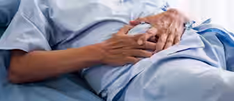 A person lying on a hospital bed wearing a light blue medical gown, with hands resting over their abdomen. The scene suggests a hospital or clinical setting with natural light filtering in from the background.