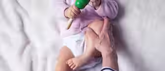 A baby in a diaper and light pink sweater holds a green maraca while sitting on a soft, plush white surface. An adult, wearing a striped shirt, gently holds the baby's legs with both hands. Only the lower legs of the adult and the baby’s hands and legs are visible.