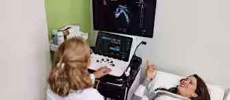A pregnant woman lies on an examination bed smiling while a healthcare professional conducts an ultrasound. The monitor shows the ultrasound image, and the room has medical equipment and supplies in the background.