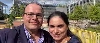 A man and a woman are smiling at the camera, appearing to be outdoors in a park or campus setting with modern buildings and trees in the background. It is a partly cloudy day. Both are dressed in casual business attire and seem to be enjoying the moment.