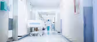 A bright hospital corridor with white walls and floors. Medical staff in blue scrubs are seen in the distance, alongside medical equipment and patient beds. The corridor is tidy, with various medical supplies and wall fixtures visible on the sides.