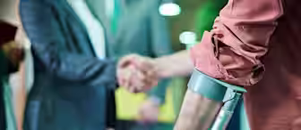 A person in a pink shirt with rolled sleeves using crutches is in the foreground. In the background, two people are shaking hands, one in a grey suit and another in a suit jacket. The scene appears to be in a professional setting with soft lighting.