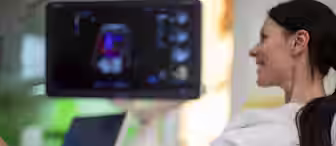 A woman is lying down, smiling during an ultrasound examination. A medical professional is using a handheld device on her abdomen. A monitor displays the ultrasound image in the background.