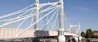 A historic suspension bridge with ornate white and pink detailing spans a broad river under a clear blue sky. The bridge has multiple cables and decorative towers, with buildings and greenery visible on the far shore.