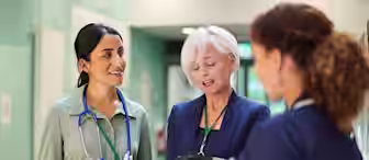 Three healthcare professionals stand in a hospital corridor, engaged in conversation. One has a stethoscope around her neck, another holds a clipboard, and the third is dressed in a nurse's uniform, holding a tablet.