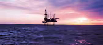 An offshore oil rig stands in the middle of a vast ocean under a dramatic sky at either sunrise or sunset. The sky is filled with a mix of purple, pink, orange, and blue hues, casting reflective colors on the water's surface.