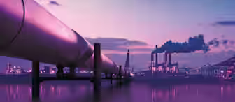 A large oil pipeline extends across a body of water towards an industrial complex with smokestacks emitting smoke. The sky is at dusk, painted in shades of purple and pink, with the water reflecting the colors and lights of the structures.