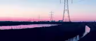 A series of electrical transmission towers and power lines stretch across a reflective water body at sunset. The sky transitions from a soft pink near the horizon to a serene blue higher up, with silhouettes of buildings and trees in the distant background.