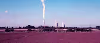 A power plant with three large cooling towers and one tall chimney emitting white smoke stands in the middle of a barren field. The sky is clear blue, and the scene is bordered by a few trees at the edge of the field.