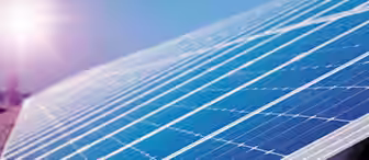 A close-up view of a large solar panel array under a clear blue sky with the sun shining brightly in the background. The solar panels reflect the sunlight, highlighting their grid-like structure and sleek surface.