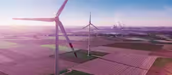 Aerial view of two large wind turbines standing in a vast, open field with a clear blue sky. Farmland stretches out in neat, colorful plots below, while industrial structures with smokestacks are visible on the horizon.