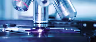 Close-up view of a microscope's objective lenses with detailed engravings visible, showing magnification levels. The image captures part of the slide stage and focus mechanisms. The background is softly blurred, focusing attention on the microscope.