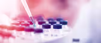 A close-up image shows a scientist with blurred facial features in the background using a pipette to place a blue liquid into one of several small, capped vials arranged in a circular holder. The scene suggests a laboratory setting.