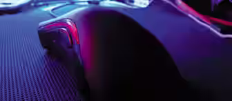 Close-up of a computer mouse on a dark mouse pad, illuminated by a purple and blue light. The background is slightly blurred but features a clutter of wires and computer components, with various LEDs and screens creating a moody, tech-centric atmosphere.