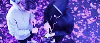 A group of four people wearing athletic wear, standing in a circle on a leaf-covered ground. They are all looking at a smartphone held by one person dressed in a hooded sweatshirt. The scene is lit with a purple hue, and they appear to be discussing something.