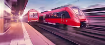 Two red trains are captured in motion on adjacent tracks, with a blurred effect that emphasizes their speed. The platform to the left is partially visible, showing empty space and overhead lights. The scene is set during twilight, with the sky transitioning to evening.