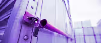 Close-up of a padlock latching a metal door, with the focus on the locking mechanism. The image has a strong purple hue, giving it a surreal, monochromatic look. Stacked containers are visible in the blurry background, suggesting an industrial setting.