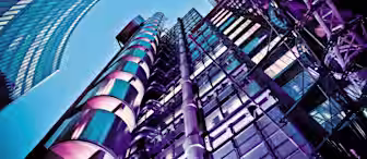 A dramatic low-angle shot of futuristic skyscrapers against a clear blue sky. The buildings, featuring sleek glass facades and metallic structures, are illuminated with vibrant purple and blue lights, emphasizing their modern architectural designs.