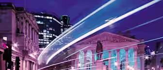 A nighttime city scene with light trails from passing vehicles, illuminating a historical building with columns and a statue in front. The sky is a deep blue, and the surrounding modern buildings are lit up, contrasting the neoclassical architecture.