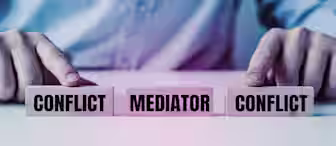 A person in a suit is seen from the torso down, pointing at three wooden blocks on a table. The blocks are labeled "CONFLICT," "MEDIATOR," and "CONFLICT." The image symbolizes the role of a mediator in resolving conflicts between two parties.