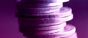 A stack of coins precariously balanced, viewed up close and illuminated with dramatic blue and purple lighting, creating a vivid contrast and giving the coins a metallic sheen. The background is blurred, keeping the focus on the coins.