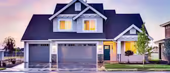 A modern two-story house with white and blue exterior, three-car garage, and a teal front door. Warm interior lights are on, and a small tree is planted in the front yard. The sky is colorful, indicating either sunrise or sunset. The driveway is slightly wet.