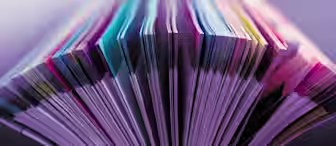 Close-up of the colorful, curved pages of an open book, fanned out to create a visually striking pattern. The pages feature various shades and hues, creating a vibrant and abstract appearance. The background is blurred in soft purple tones.