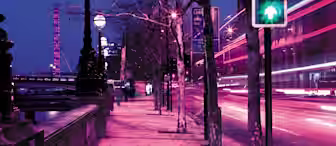 A night scene of a city street with motion blur effects from passing vehicles. The pavement is lit by street lamps. Light trails from the vehicles create a dynamic atmosphere. The traffic light is green, and bare trees line the sidewalk.