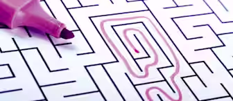 A close-up of a maze puzzle partially solved with a pink highlighter. The highlighter is at the edge of the maze, which has a thick, pink line marking a winding path from the center towards the top left corner.