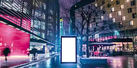 A brightly lit, modern urban plaza at night with buildings on both sides. In the center, there is a large blank digital billboard. People are walking around, and a few trees and outdoor seating areas are visible. The ground is wet, reflecting the lights.