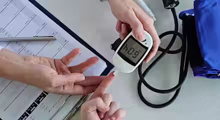 A healthcare professional uses a glucometer to check a patient's blood sugar level, displaying 6.09. A blood pressure monitor, pen, and medical chart are on the table nearby.