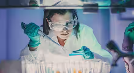 A scientist wearing a lab coat, safety goggles, and protective gloves is using a pipette to transfer liquid into a petri dish. She is surrounded by test tubes filled with colorful liquids and appears focused on her work in a laboratory setting.