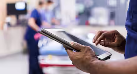 A close-up of a healthcare worker’s hands holding and using a digital tablet in a hospital setting, with medical staff and equipment blurred in the background.