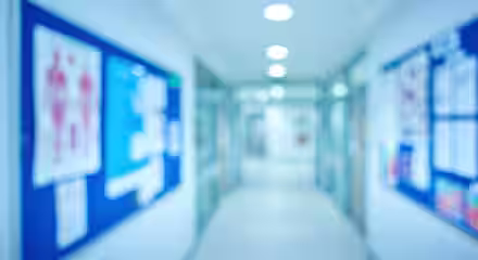 A blurred image of a hospital or clinic hallway with bright overhead lighting. Blue and white informational boards displaying various posters and charts line the walls. The floor and walls are white, contributing to a sterile and clinical environment.