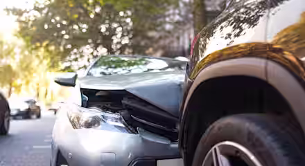 A silver car with a damaged front bumper has collided with the rear side of a black SUV on a city street lined with trees.
