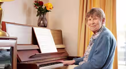 An elderly woman with short gray hair and wearing a blue sweater sits at a piano with sheet music. She smiles gently at the camera. A vase with a yellow and red flower arrangement is placed on the piano, and yellow curtains hang in the background.