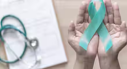 Hands holding a teal ribbon over a clipboard with a stethoscope, symbolizing ovarian cancer awareness. The background is a wooden surface.