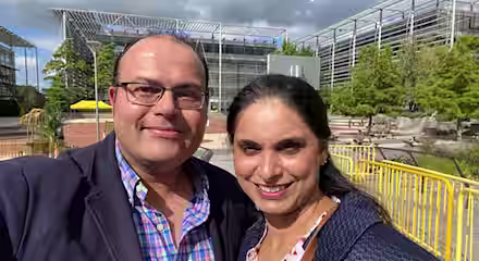 A man and a woman are smiling at the camera, appearing to be outdoors in a park or campus setting with modern buildings and trees in the background. It is a partly cloudy day. Both are dressed in casual business attire and seem to be enjoying the moment.
