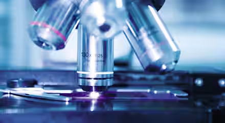 Close-up view of a microscope's objective lenses with detailed engravings visible, showing magnification levels. The image captures part of the slide stage and focus mechanisms. The background is softly blurred, focusing attention on the microscope.