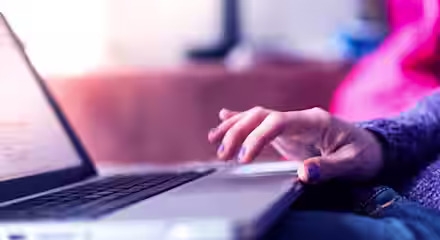 Close-up of a person's hand with purple nail polish resting on the trackpad of an open laptop. The background is softly blurred with pink and blue tones. The person appears to be using the laptop, which is situated on a flat surface.