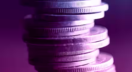 A stack of coins precariously balanced, viewed up close and illuminated with dramatic blue and purple lighting, creating a vivid contrast and giving the coins a metallic sheen. The background is blurred, keeping the focus on the coins.