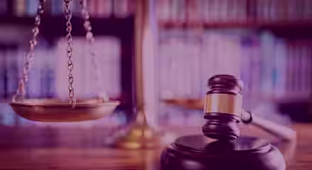 A wooden gavel rests on a sound block in the foreground, with a balanced scale of justice in the background. The setting appears to be a courtroom or a law library, indicated by the blurred shelves filled with books.