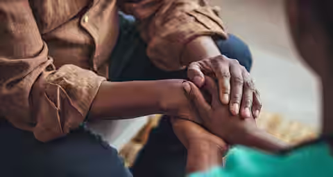 Close-up of two people holding hands, sitting across from one another. One person wears a brown shirt, and the other wears a turquoise shirt. The image captures a moment of comfort and support between them.