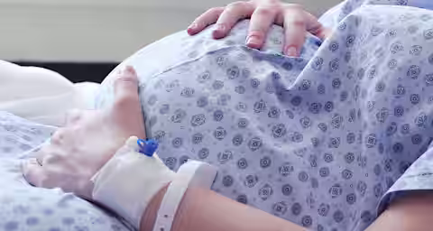 A pregnant person in a hospital bed wearing a patterned gown rests their hands on their belly. An IV is attached to their wrist, indicating they are receiving medical care.