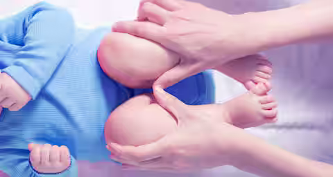 A baby wearing a blue knitted outfit lies on a soft surface, with an adult's hands gently massaging the baby's thighs. The image is focused on the legs and the hands, showing a close-up of the interaction.