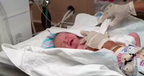 A newborn baby wrapped in a white blanket lies on a hospital bed, crying. A healthcare professional wearing gloves and a colorful scrub top is examining and tending to the baby. Medical equipment is visible in the background.